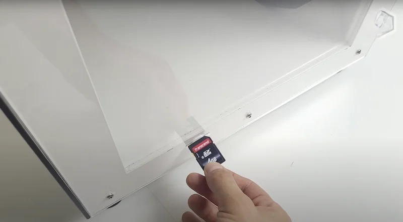 Hand inserting an SD card into an Anisoprint Composer A3 3D printer, showcasing its sleek white design and clear panel.