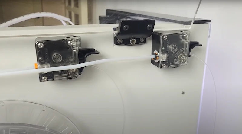 Anisoprint Composer A3 printer showing filament feeding mechanism with two stepper motors and clear tubing against a white background.