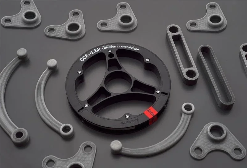 Anisoprint Composer A3 showcases composite carbon fiber parts on a gray background, featuring gears and structural components.