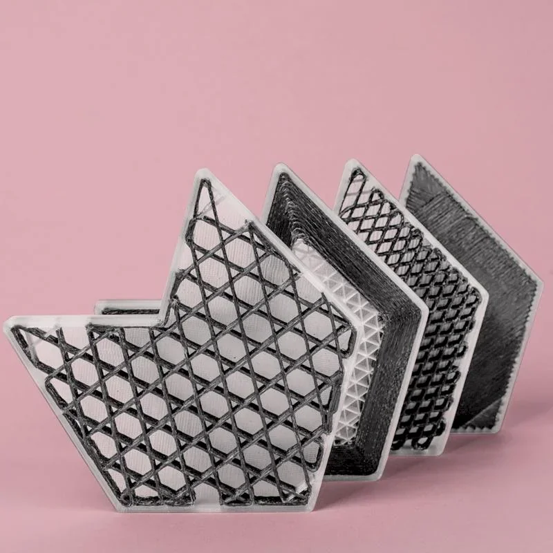 3D-printed geometric parts with lattice designs created by anisoprint composer a3, set against a pink background.
