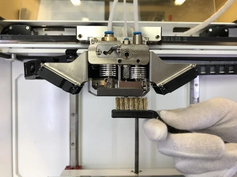 Anisoprint Composer A4 extruder close-up with gloved hand cleaning brass brush tool, set in a workshop environment.