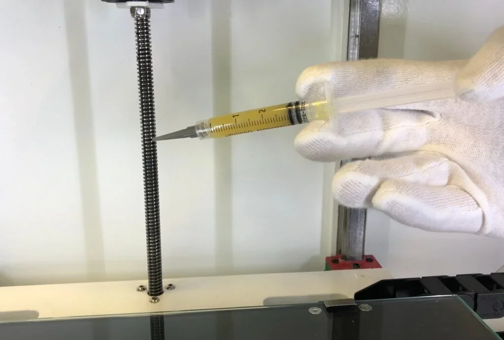 Gloved hand holding syringe near a metal spring inside an Anisoprint Composer A4 3D printer, demonstrating maintenance or calibration process.