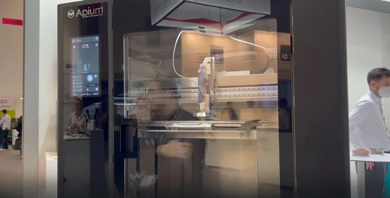 Apium P400 3D printer with transparent casing shown at an exhibition, creating a detailed part, visible control panel and branding.