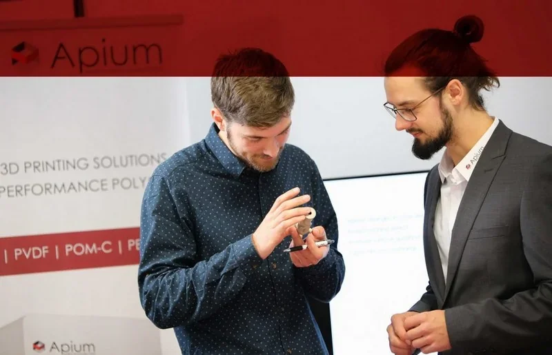 Two men examine a 3D-printed part, likely made by the Apium P400, against a branded backdrop at a trade show or presentation.