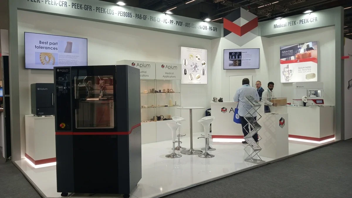 Apium P400 3D printer showcased in a trade booth with samples, surrounded by displays and branding, highlighting industrial and medical applications.