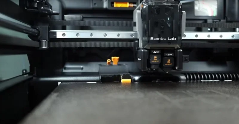 Bambu Lab H2D 3D printer, close-up of print head inside, with detailed view of rails and small sample parts created by the printer.