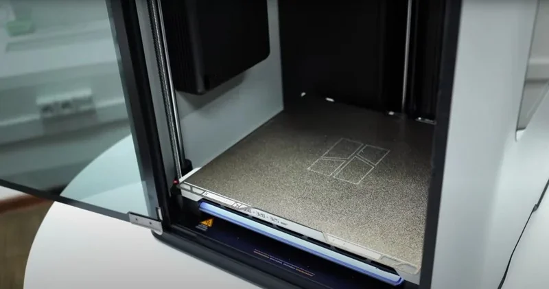 Bambu H2D 3D printer with open chamber, showing metallic print bed and sample structures created by the printer, situated in a clean workspace.