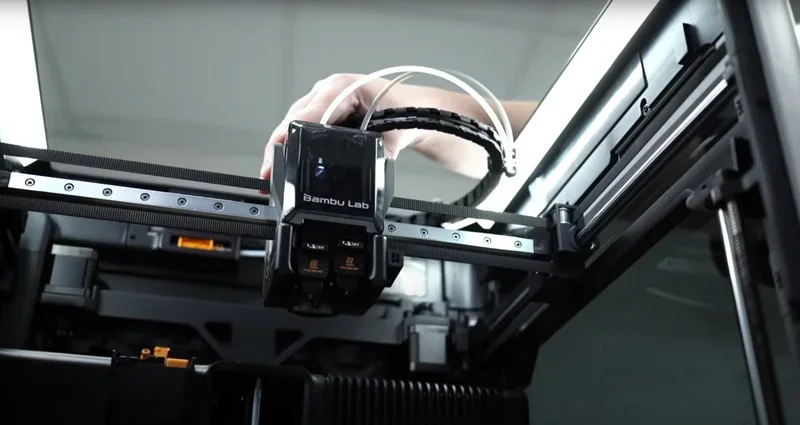 Bambu Lab 3D printer close-up, hand adjusting the Bambu H2D extruder mechanism inside a sleek, modern enclosure.