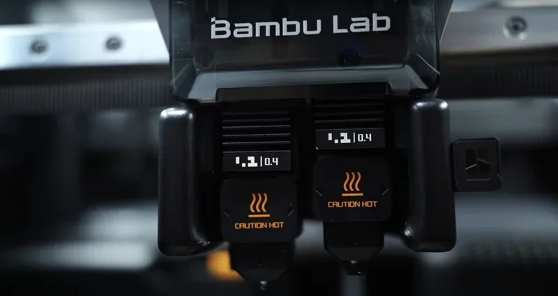 Bambu Lab H2D 3D printer nozzle close-up, showing two hot nozzles with "Caution Hot" labels, in a sleek industrial design.