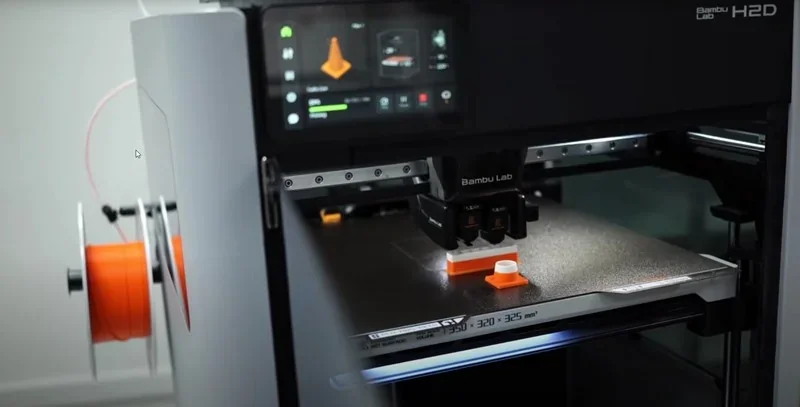 Bambu H2D 3D printer creating orange and white parts, with filament spool, control screen displaying settings, in a modern workspace.