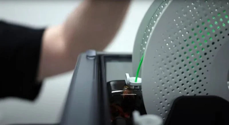 Bambu H2D 3D printer with filament spool, arm adjusting spool, creating parts. Sleek design, modern tech, focus on filament feed mechanism.