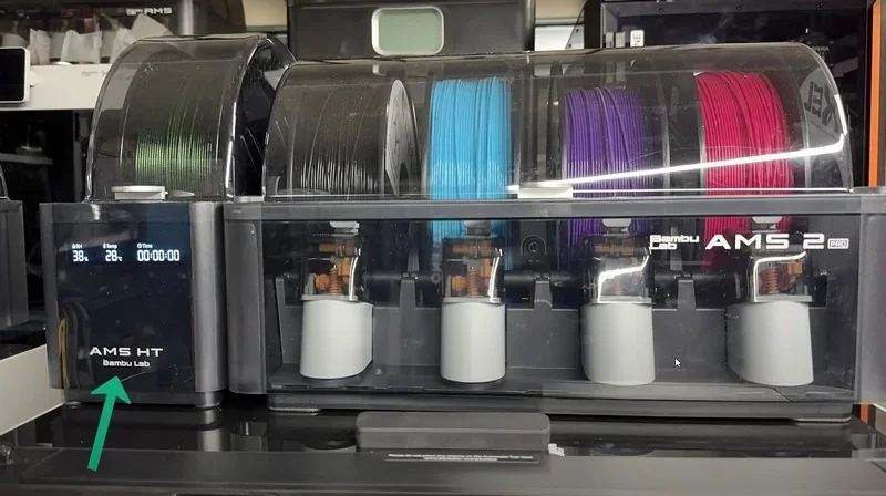 Bambu H2D 3D printer with AMS 2 Pro, displaying multiple filament spools in black, blue, purple, and pink colors, branding visible on device.
