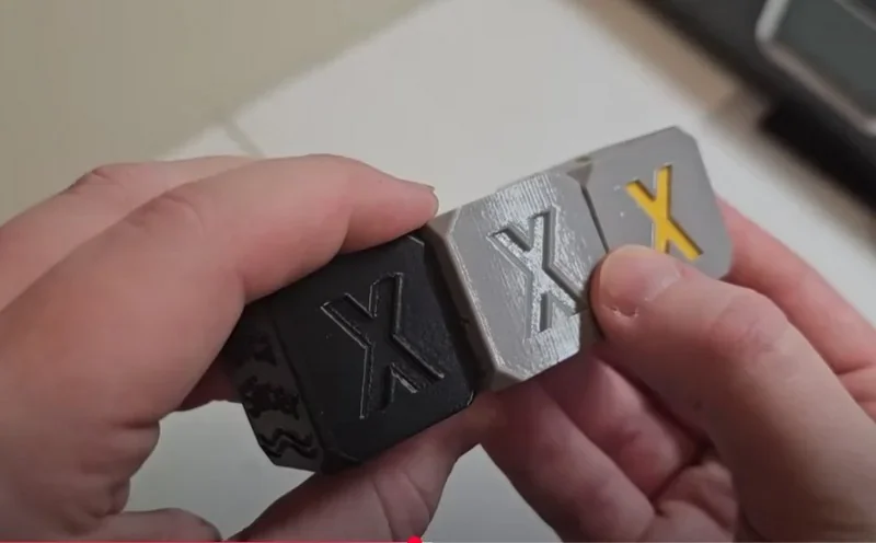 Hands holding black, gray, and white 3D-printed calibration cubes with "X" from a Bambu H2D printer.