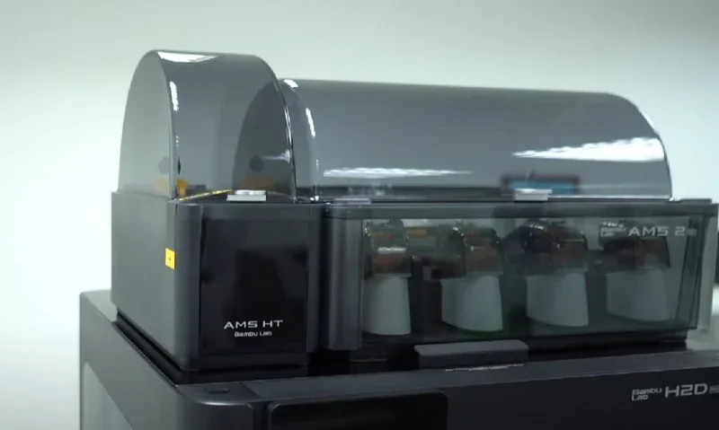 Bambu Lab H2D Pro 3D printer featuring AMS HT module with filament spools, sleek gray design, and visible branding in a bright indoor setting.