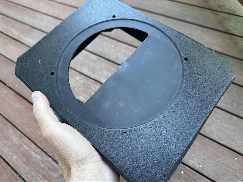 Black 3D-printed part with circular cutout held over wooden deck, created by bambu lab h2d pro.