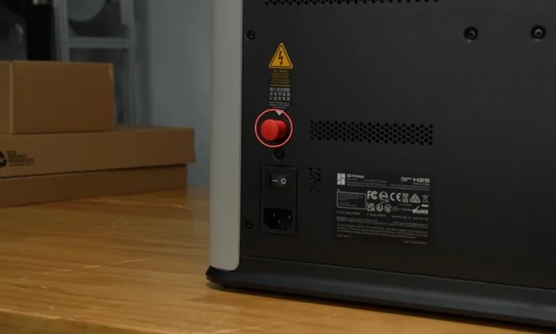 Bambu Lab H2S 3D printer back panel with power switch, red reset button, and safety labels on a wooden table, boxes in the background.