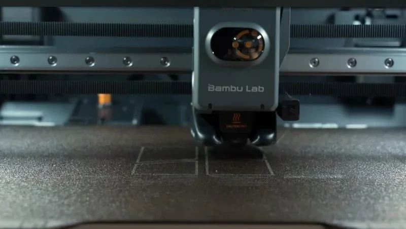 Bambu Lab H2S 3D printer close-up, printing precise outlines on a textured surface; branding visible on the machine.