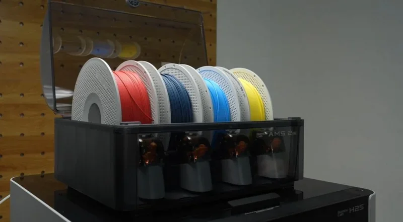 Bambu Lab H2S 3D printer with multiple filament colors: red, black, blue, and yellow on spools, set on a pegboard backdrop.