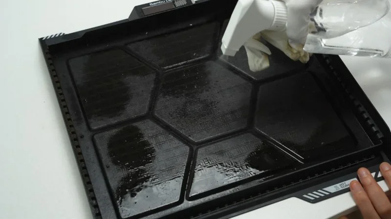 Bambu Lab H2S print tray being cleaned with a spray bottle and glove. Hexagonal pattern visible on the tray surface, showcasing care process.