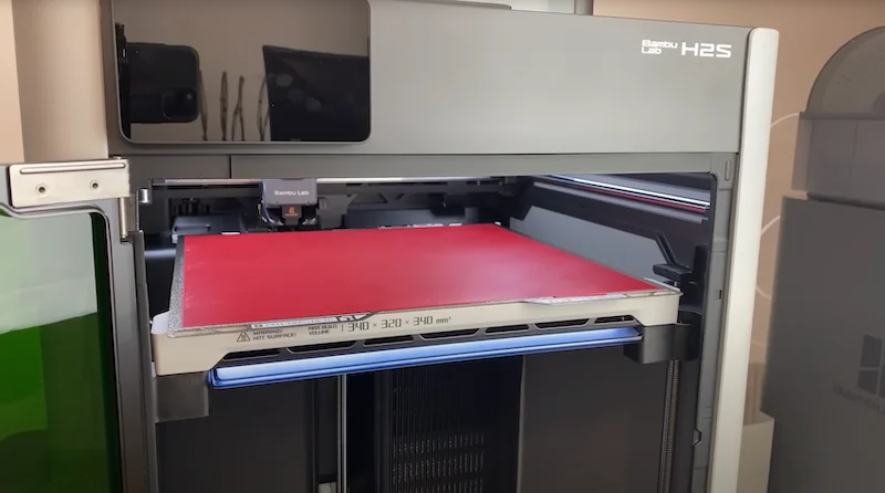 Bambu Lab H2S 3D printer with open tray, red print bed, sleek design, visible branding; high-tech equipment in a modern setting.