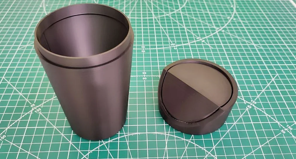 3D-printed black cup and lid on green grid mat, crafted by Bambu Lab X1.