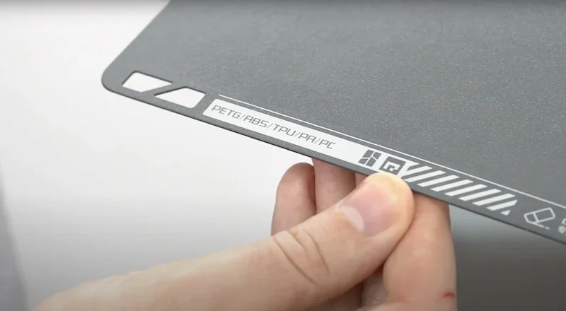 Bambu Lab X1 print bed held by hand, labeled for PETG, ABS, TPU, PA, PC materials, with branding icons and gray surface texture visible.