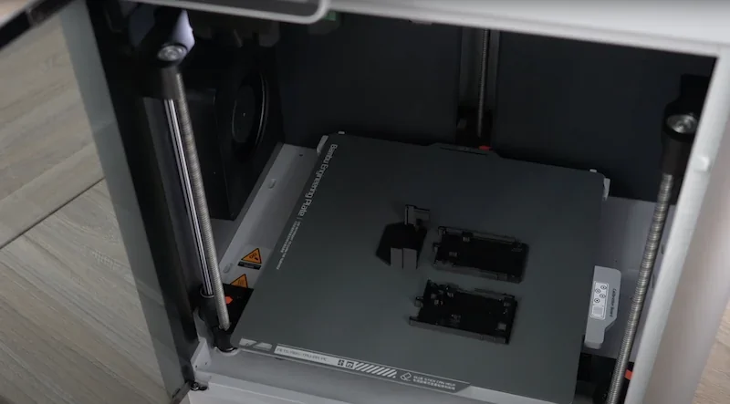 Bambu Lab X1 3D printer interior showing printing bed with two black sample parts, enclosed in a sleek metallic case, highlighting precision engineering.