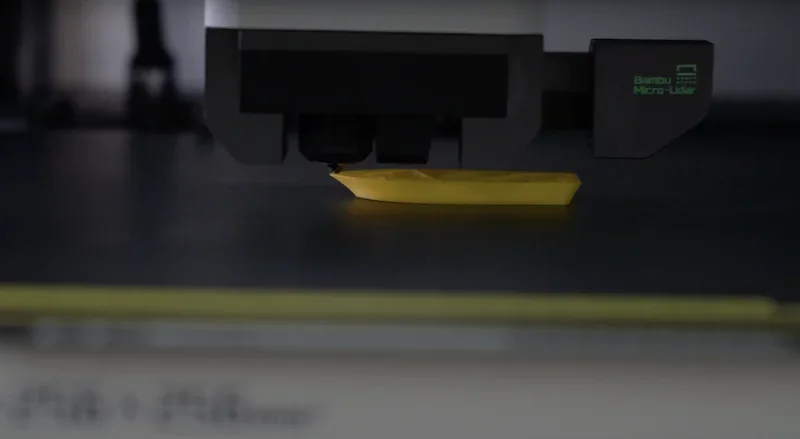 Bambu Lab X1 printing yellow 3D part, close-up of extruder head with branding visible, focused on precision and technology.