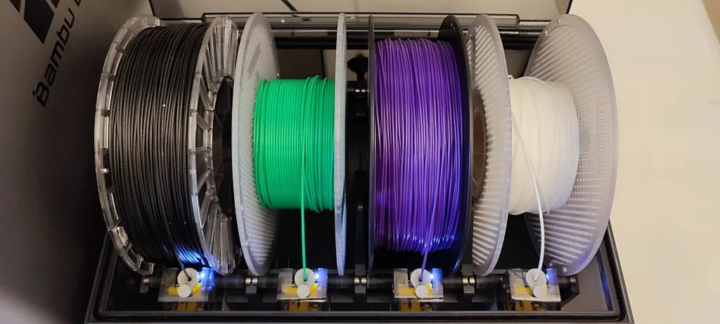 Bambu Lab X1 3D printer AMS unit with black, green, purple, and white filament spools, top view, inside branded enclosure, four extruders visible.