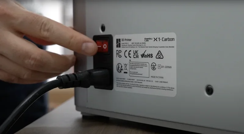 Bambu Lab X1 Carbon power switch and certification labels, hand toggling the switch, emphasizing the 3D printer's technical details and branding.