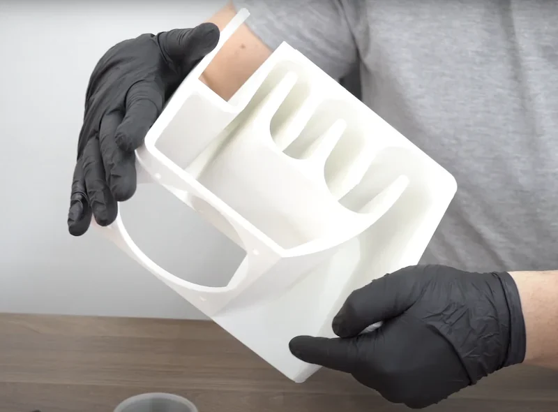 Hands in black gloves hold a 3D-printed white object created by Bambu P1P, featuring compartments and spiral design, on a wooden table.