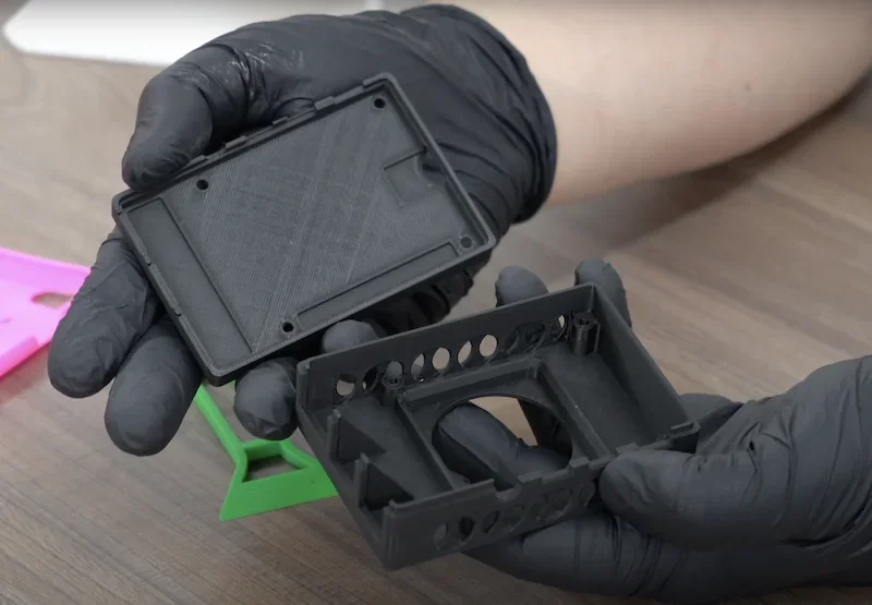 3D-printed parts created by bambu p1p, held by gloved hands over a wooden surface, with colorful printed samples in the background.