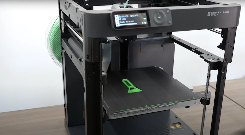 Bambu Lab P1P 3D printer with a black frame, digital display, and a partially printed green object, set on a wooden surface.