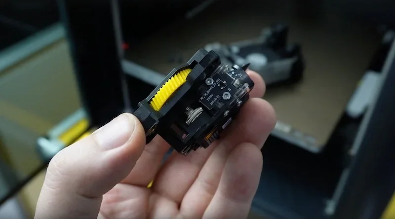 Hand holding a 3D printed component with gears created by a Bambu P1P printer, with the printer partially visible in the background.