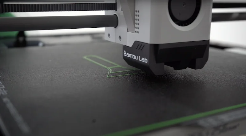 Bambu P1P 3D printer in action, nozzle over the print bed laying green filament, featuring Bambu Lab branding.