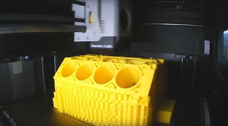 Bambu P1S 3D printer crafting a yellow engine block model in a dimly lit setting, showcasing detailed structure and precision.