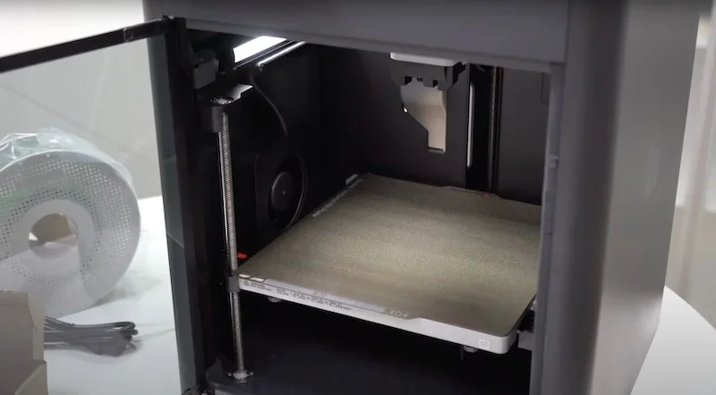 Bambu P1S 3D printer open chamber showing print bed and nozzle; filament spool visible nearby.