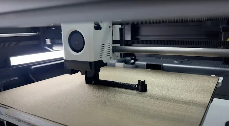 Bambu P1S 3D printer creating a black plastic part on a textured print bed, showing the print head in detail within its open enclosure.