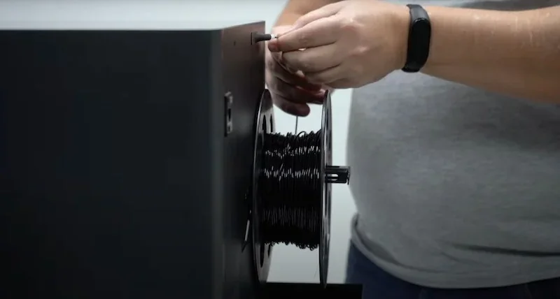 Bambu P1S 3D printer with a user loading black filament spool, showcasing the setup process. Hands, grey shirt, and watch visible, focus on the machine.