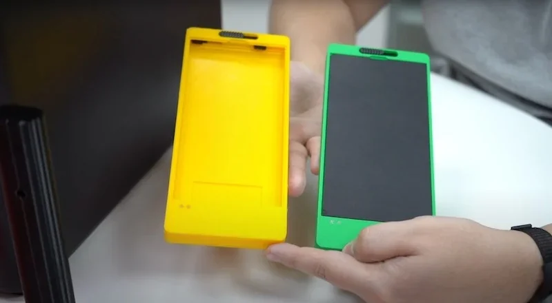 Hands holding 3D-printed yellow and green plastic phone cases by Bambu P1S on a desk, showcasing product capabilities and detailing.