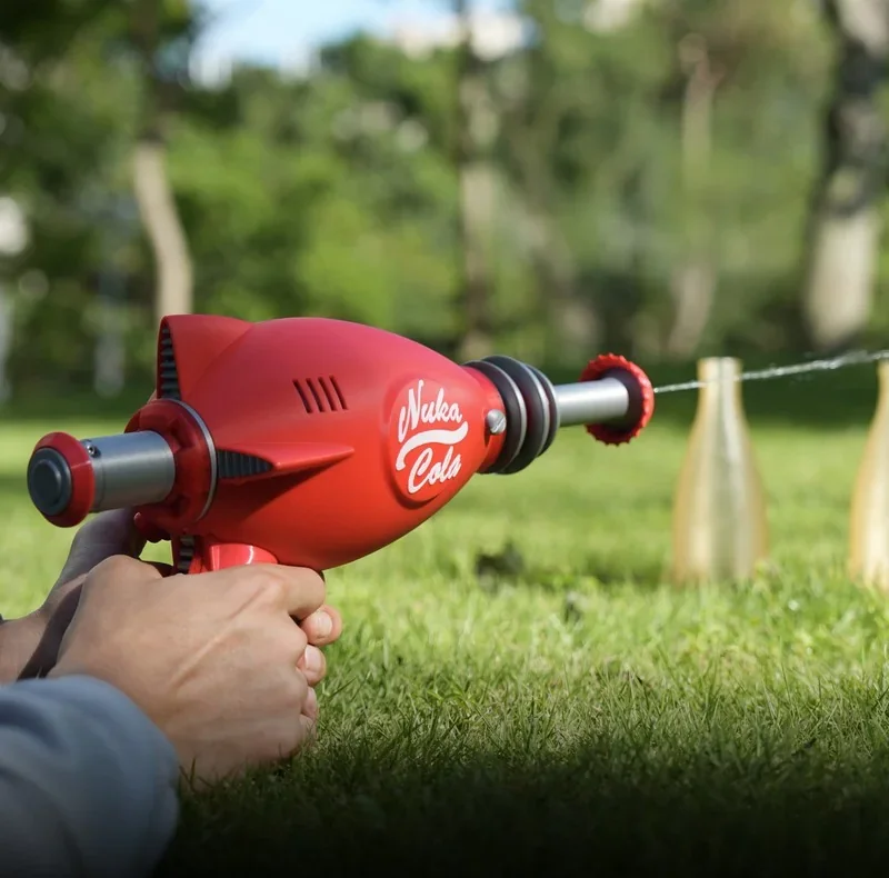 Red plastic water gun labeled “Nuka Cola,” held outdoors, aimed at bottle targets, 3D printed by Bambu P2S, on a grassy lawn.