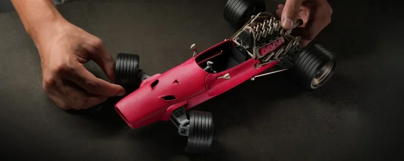 Red model race car being assembled by hand, showcasing detail created by Bambu P2S 3D printer; features intricate engine and tire components.
