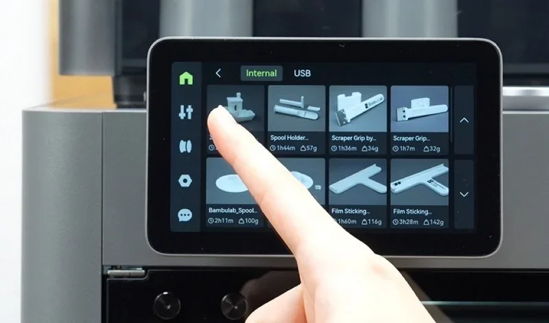 Finger using Bambu P2S 3D printer touchscreen, displaying parts like spool holders and scrapers created by the printer, on a sleek gray casing.