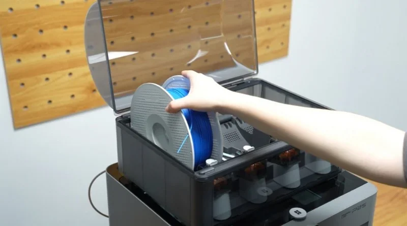 A person loads blue filament into the Bambu P2S 3D printer with a pegboard wall in the background.