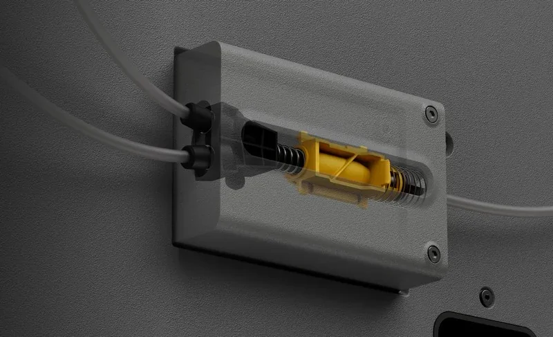 3D printer component with clear casing and yellow part, likely from Bambu P2S, showing internal mechanisms and dual tubing connections on a gray surface.