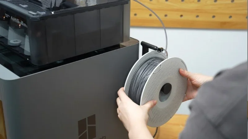 Bambu P2S 3D printer beside filament spool being loaded, showcasing design and branding in a workshop setting.