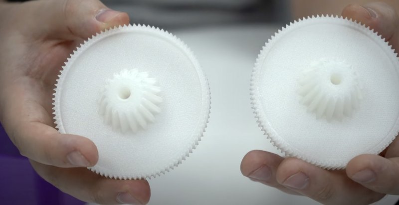 Two white, intricately designed gears are held, showcasing the precision and detail attainable with the Bambu Lab X1E 3D printer. The gears have a fine-tooth structure, highlighting the printer's capability to produce complex mechanical components. The background is softly blurred, focusing attention on the craftsmanship of the printed parts.
