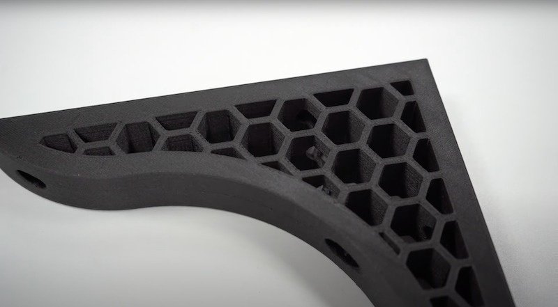 A black 3D-printed part created by the Bambu X1E, featuring a complex honeycomb internal structure for strength and reduced weight. The part, a corner bracket, appears robust and durable, with smooth curves and precise edges. The detailed view of the infill pattern highlights the printer's capability for intricate designs and structural integrity.