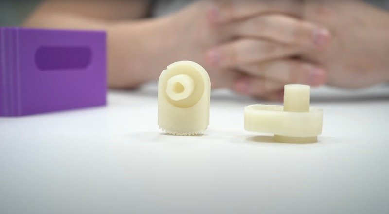 Two small, cream-colored 3D printed parts are displayed on a white tabletop. These parts, created by the Bambu X1E, showcase precise detailing and smooth surface finishes. The parts appear to be mechanical components, with one resembling a gear-like structure and the other featuring a cylindrical peg. In the background, a person's hands are resting on the table, and a purple rectangular object is visible, suggesting a context of assembly or demonstration. The focus is clearly on the quality and intricacy of the printed components.