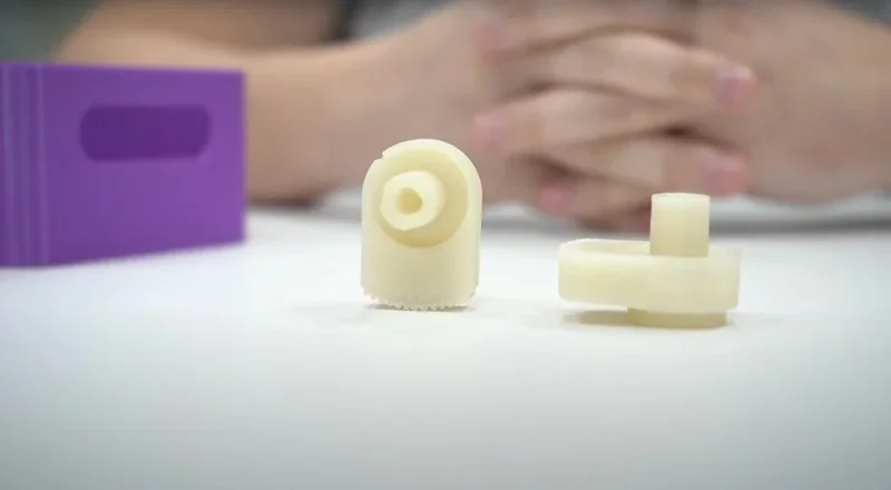 3D-printed components in beige with a purple block behind, created by the Bambu X1E printer, hands folded in the background.